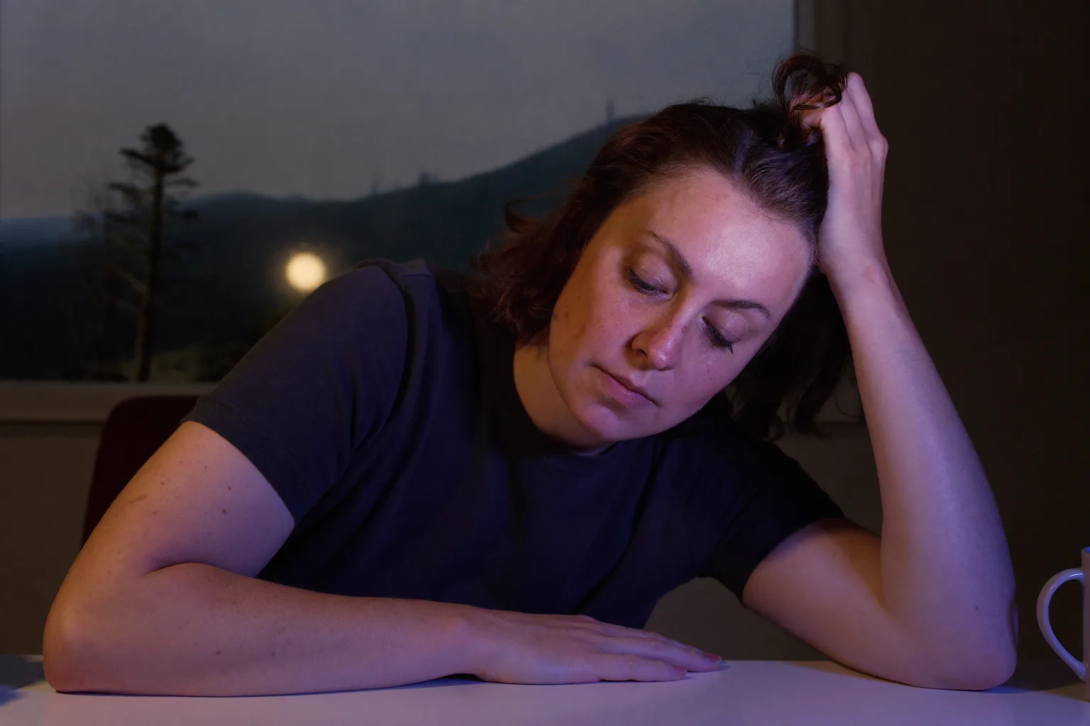 A person at a home desk late in the evening, head tilted into one hand with the fingers in their hair, eyes lowered and unfocused, the other forearm lying still on an empty desk
