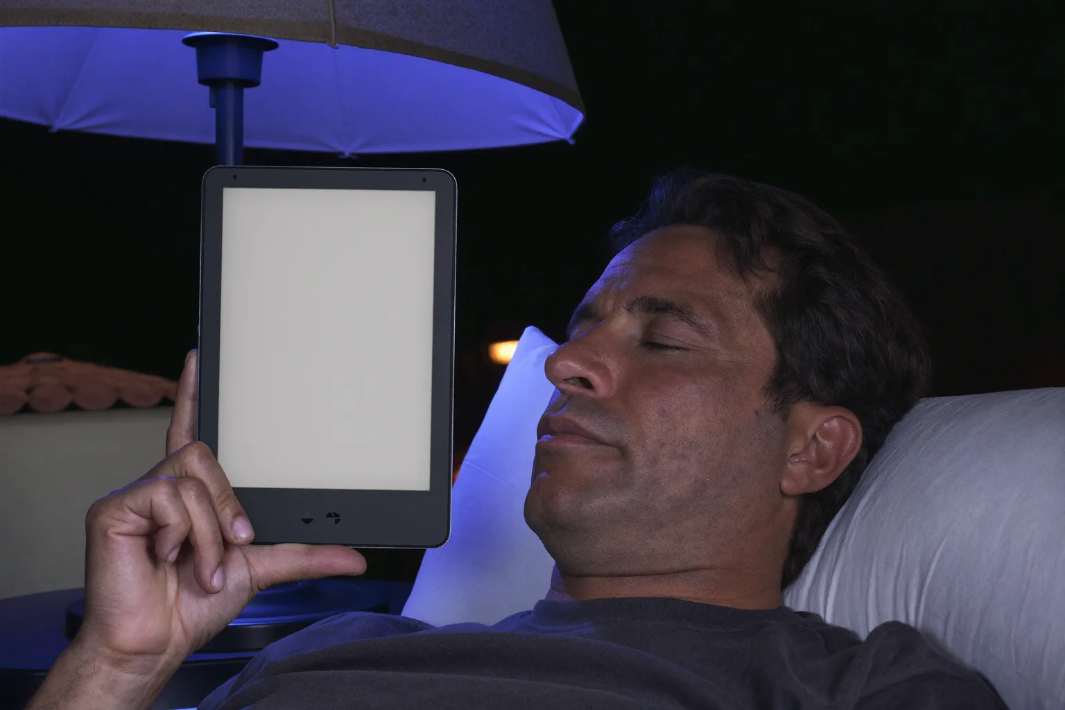 Un home llegeix en un lector electrònic al llit, ja de nit, amb la pàgina fent una resplendor tènue que amb prou feines li arriba a la cara, mentre a la tauleta de nit, al seu costat, un mòbil bocamunt inunda la tulipa del segon llum amb una llum blava freda i dura