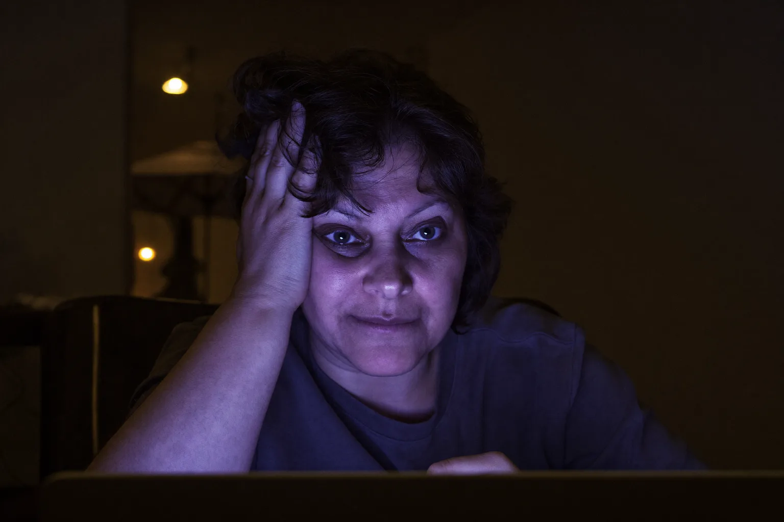 Una dona cansada davant d’un portàtil a última hora del dia, recolzant el cap a la mà, amb els ulls pesats i secs i ulleres fosques en una habitació poc il·luminada