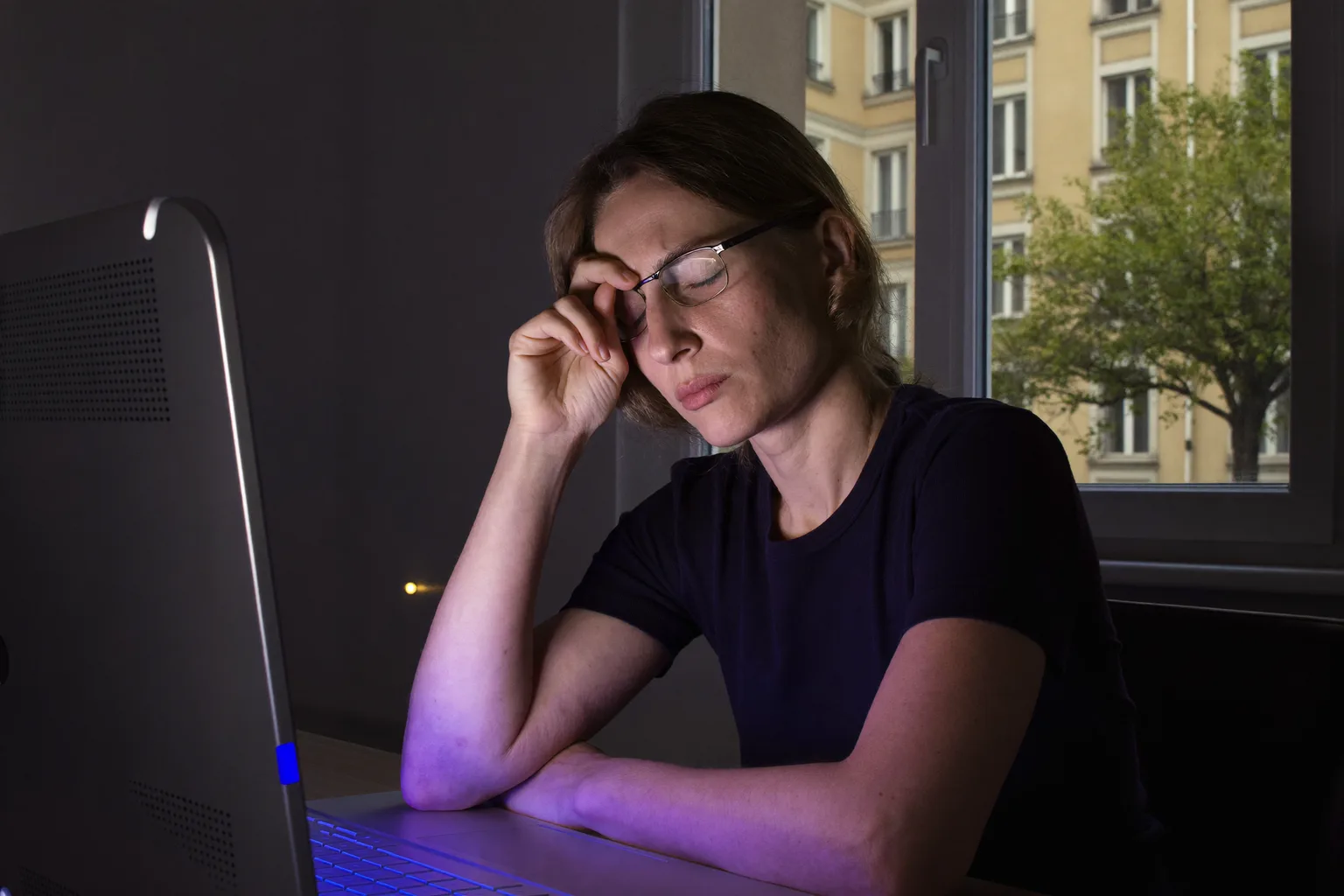 Žena u pracovního stolu v tmavé místnosti, se zavřenýma očima a brýlemi vyhrnutými na čelo, si mne kořen nosu nad zadní stranou monitoru, obličej má nasvícený studeným modrým světlem obrazovky