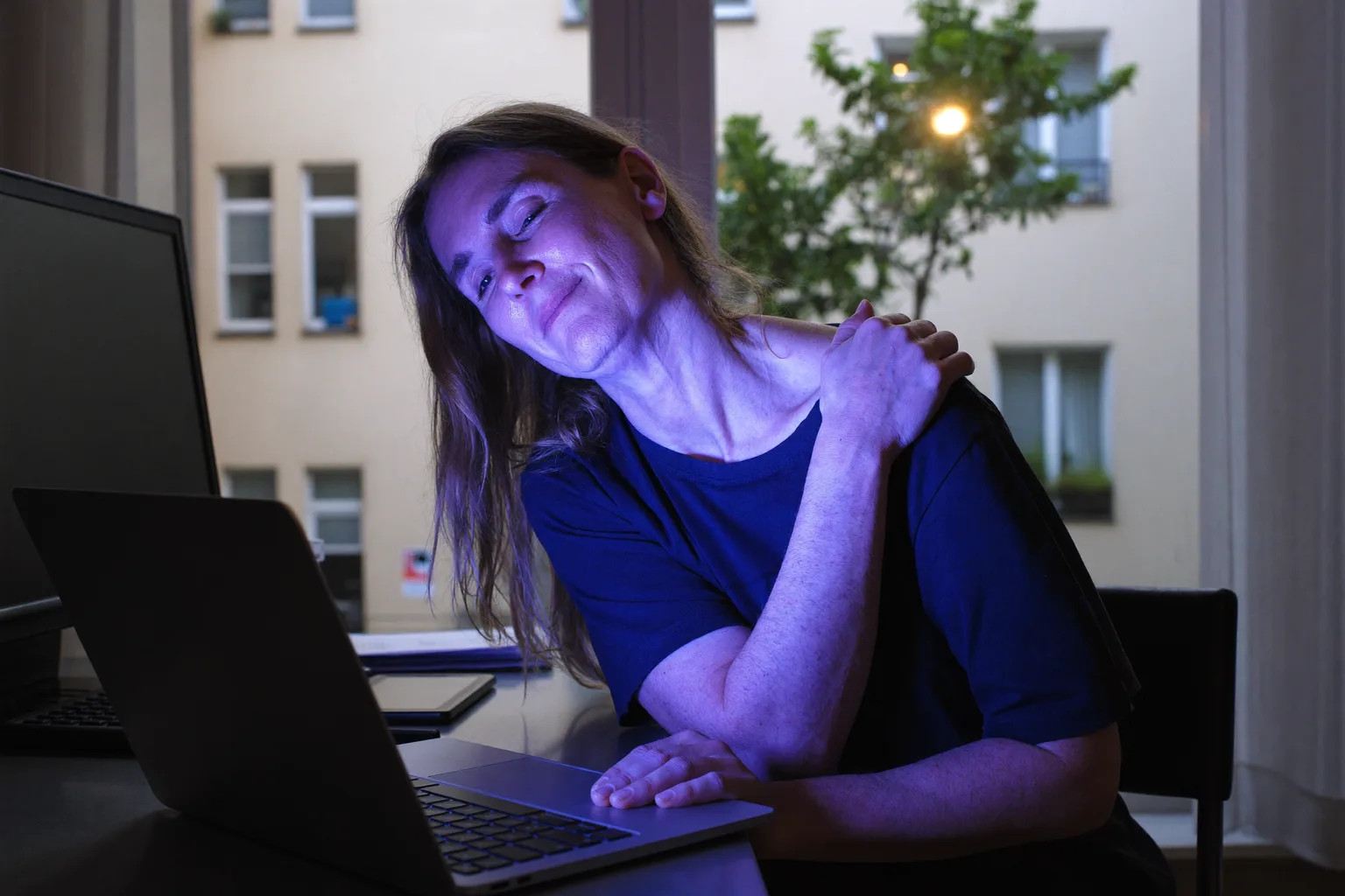 Člověk u stolu si sahá dozadu na zátylí a mne si ho před notebookem, typický obraz ztuhlého, bolavého krku po dlouhém dni u počítače