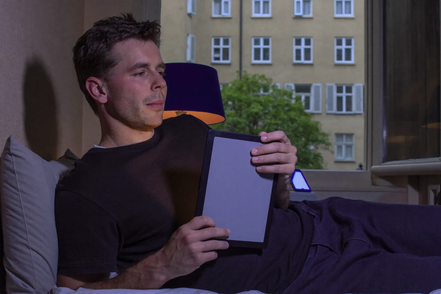 Muž čte pozdě v noci v posteli z čtečky e-knih, jejíž stránka tlumeně svítí a sotva osvětluje jeho tvář, zatímco na nočním stolku vedle něj leží telefon obrazovkou nahoru a zalévá druhé stínítko lampy tvrdým studeným modrým světlem