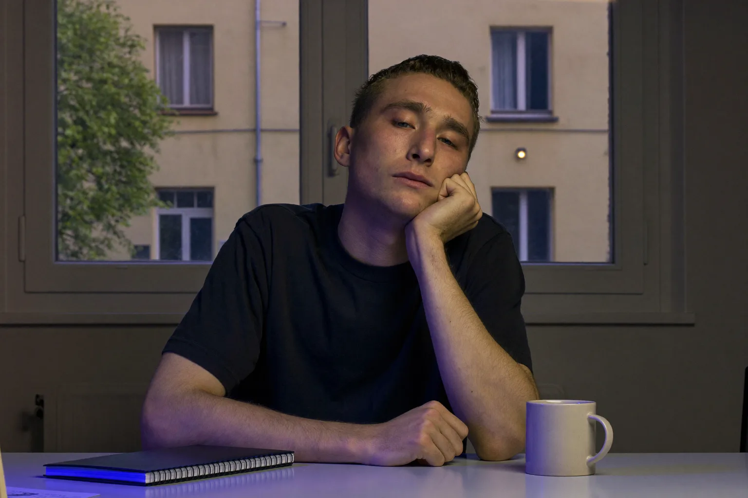 Studentka pozdě večer u stolu, bradu opřenou v dlani, tvář nasvícenou ze strany světlem obrazovky, pohled zatoulaný pryč od práce, mimo záběr