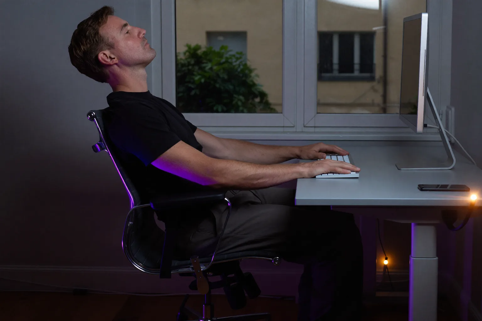 Osoba viděná z boku sedící u stolu s chodidly naplocho na podlaze, zády opřenými o opěrku židle, předloktím ve výšce stolu a monitorem v úrovni očí, vzorové držení těla u stolu