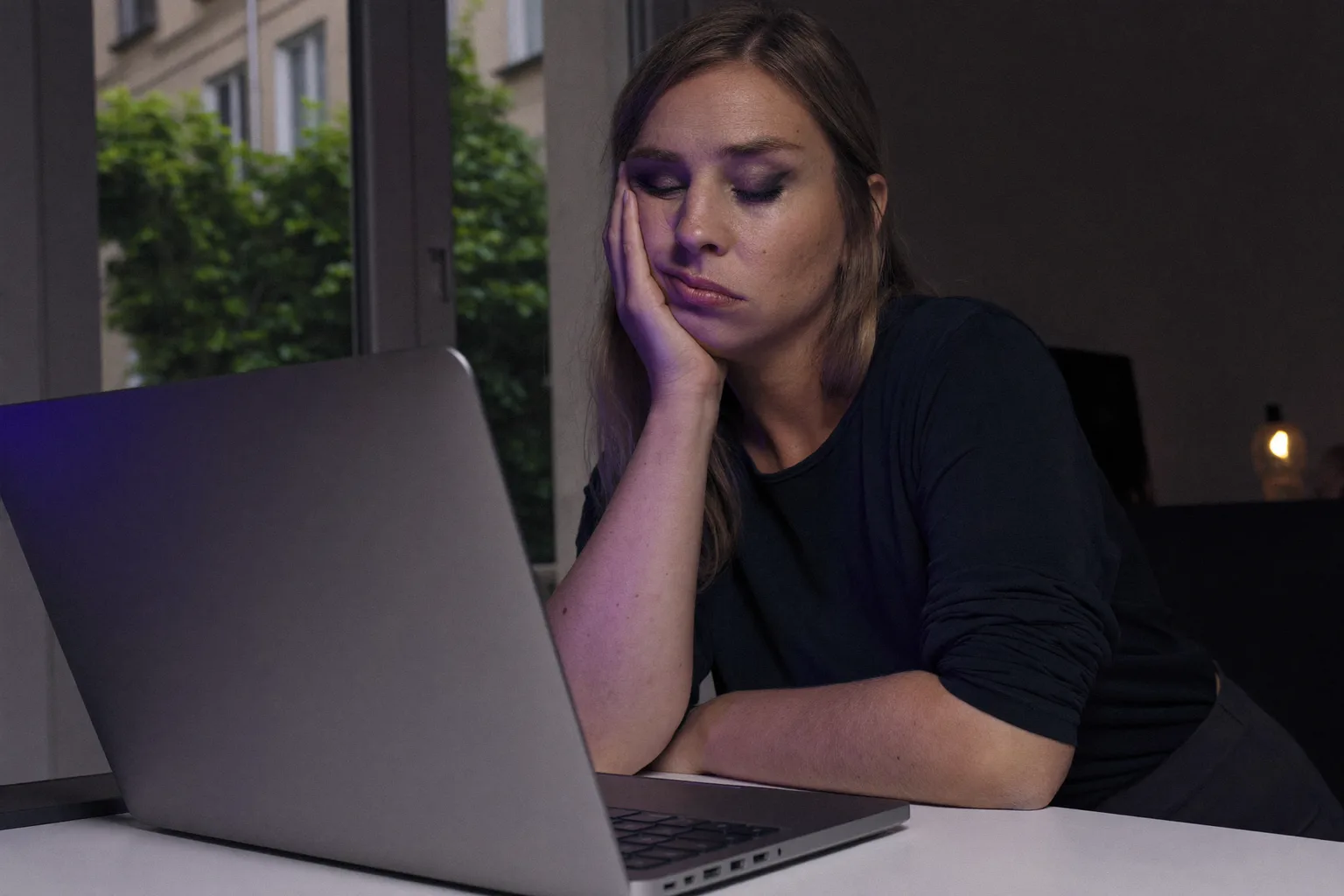 Unavená žena u notebooku pozdě večer, hlavu opřenou o dlaň, s těžkýma, suchýma očima a kruhy pod očima v tlumeně osvětlené místnosti