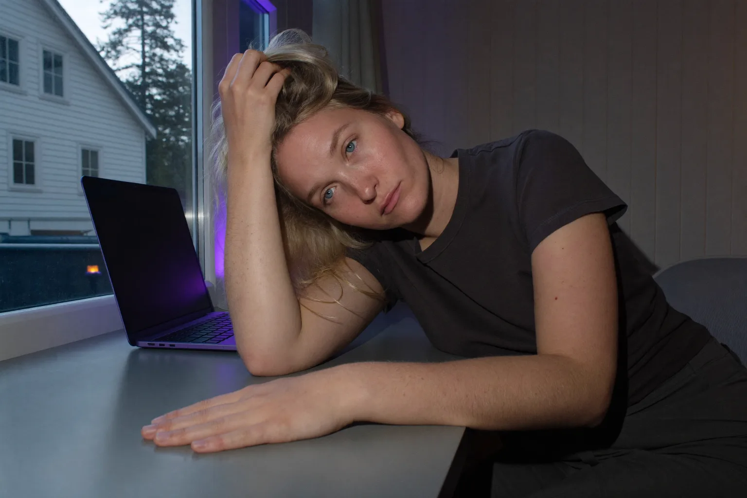 En person ved et hjemmekontor sent om aftenen, med hovedet lænet ind i den ene hånd og fingrene i håret, blikket sænket og uden fokus, mens den anden underarm ligger stille på et tomt skrivebord