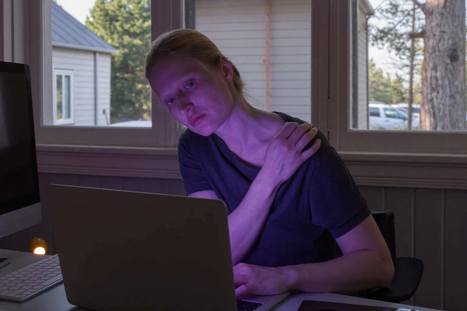En person ved et skrivebord, der rækker om bagved for at gnide kraniebasen foran en bærbar computer, og viser den stive, ømme nakke efter en lang dag ved computeren