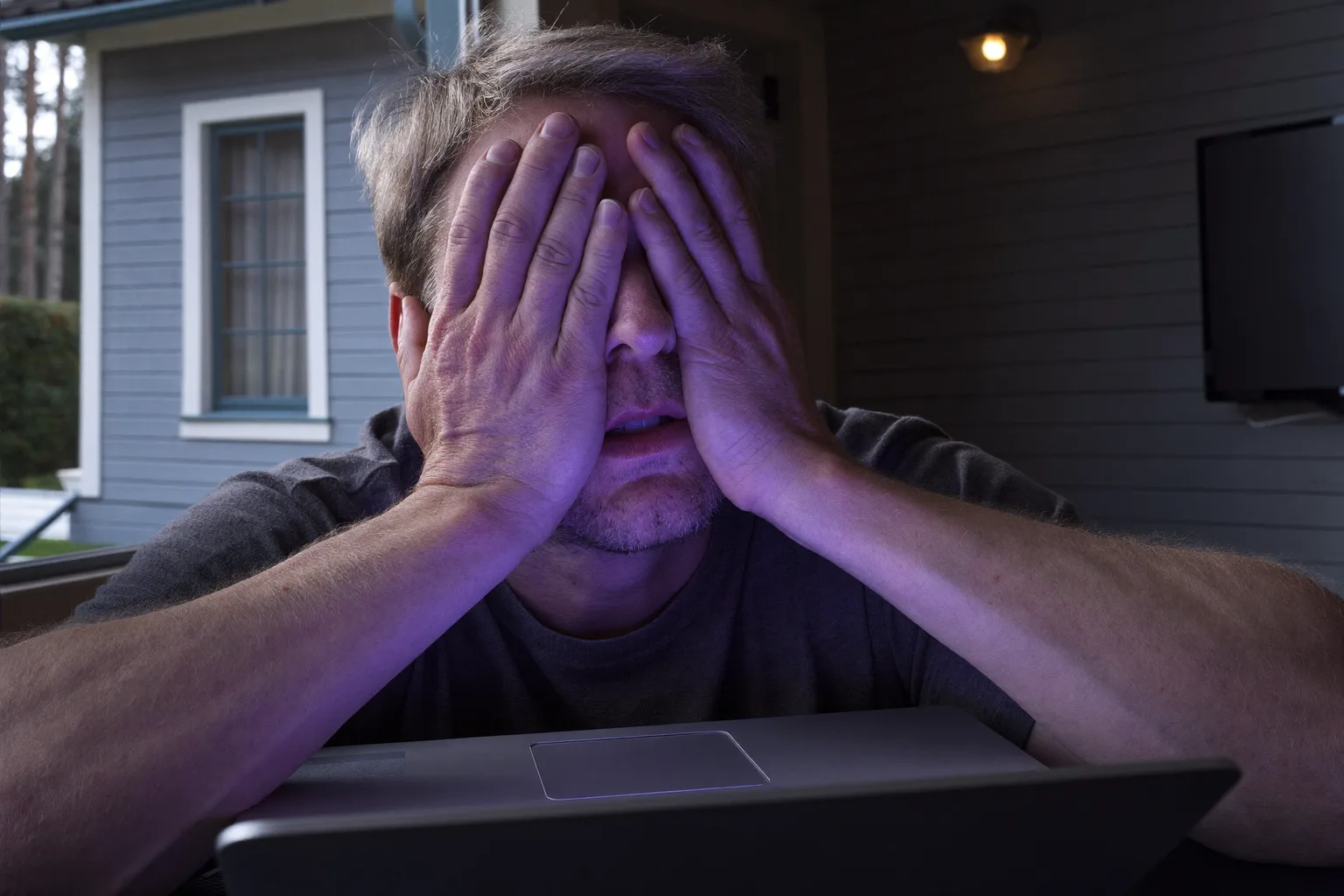 En mand ved et skrivebord i et dæmpet rum, der presser håndfladerne mod sine lukkede øjne for at lindre trætte øjne, med ansigtet oplyst af laptopskærmen