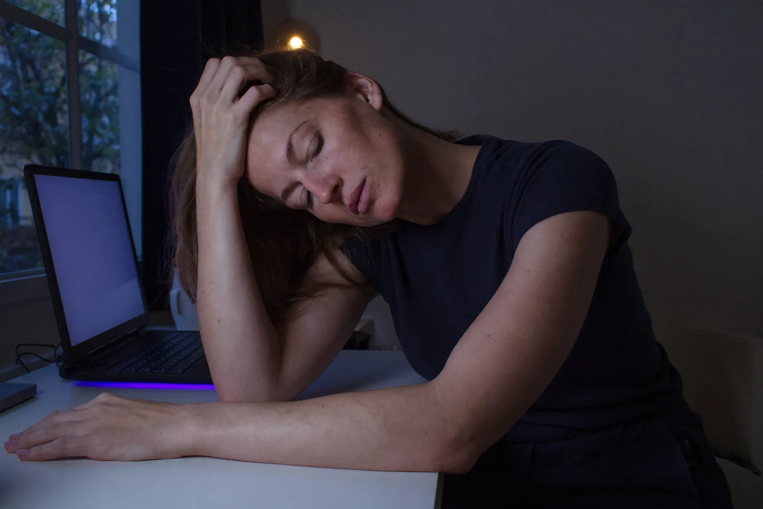 Eine Person an einem Schreibtisch zu Hause, spät am Abend, den Kopf in eine Hand gestützt, die Finger im Haar, der Blick gesenkt und unfokussiert, der andere Unterarm reglos auf dem leeren Schreibtisch