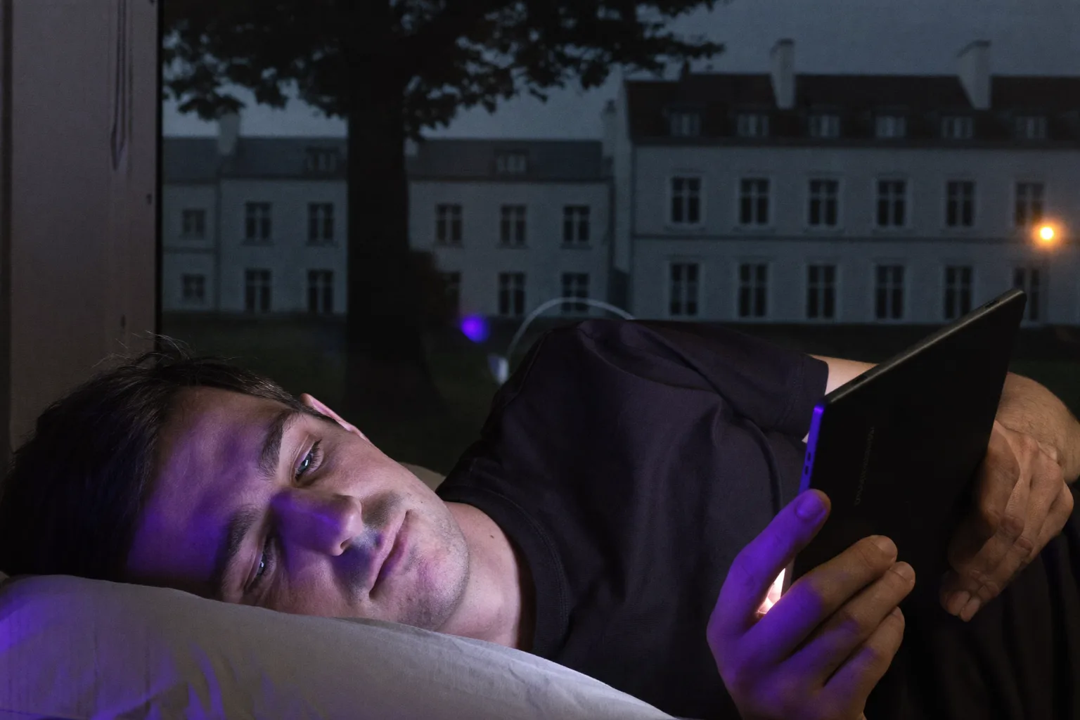 Ein Mann liest spätnachts im Bett von einem E-Reader, dessen Seite nur ein schwacher Schein ist, der sein Gesicht kaum erreicht, während auf dem Nachttisch daneben ein Handy mit dem Display nach oben den zweiten Lampenschirm in hartes, kaltes Blaulicht taucht.
