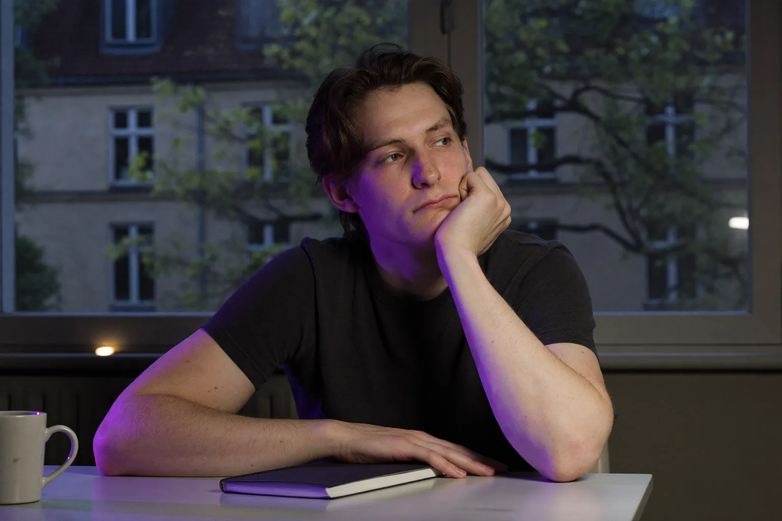 Eine Person spät am Abend an einem Schreibtisch, das Kinn in eine geöffnete Handfläche gestützt, das Gesicht seitlich vom Bildschirmlicht beleuchtet, der Blick von der Arbeit abgewandt und aus dem Bild heraus gerichtet