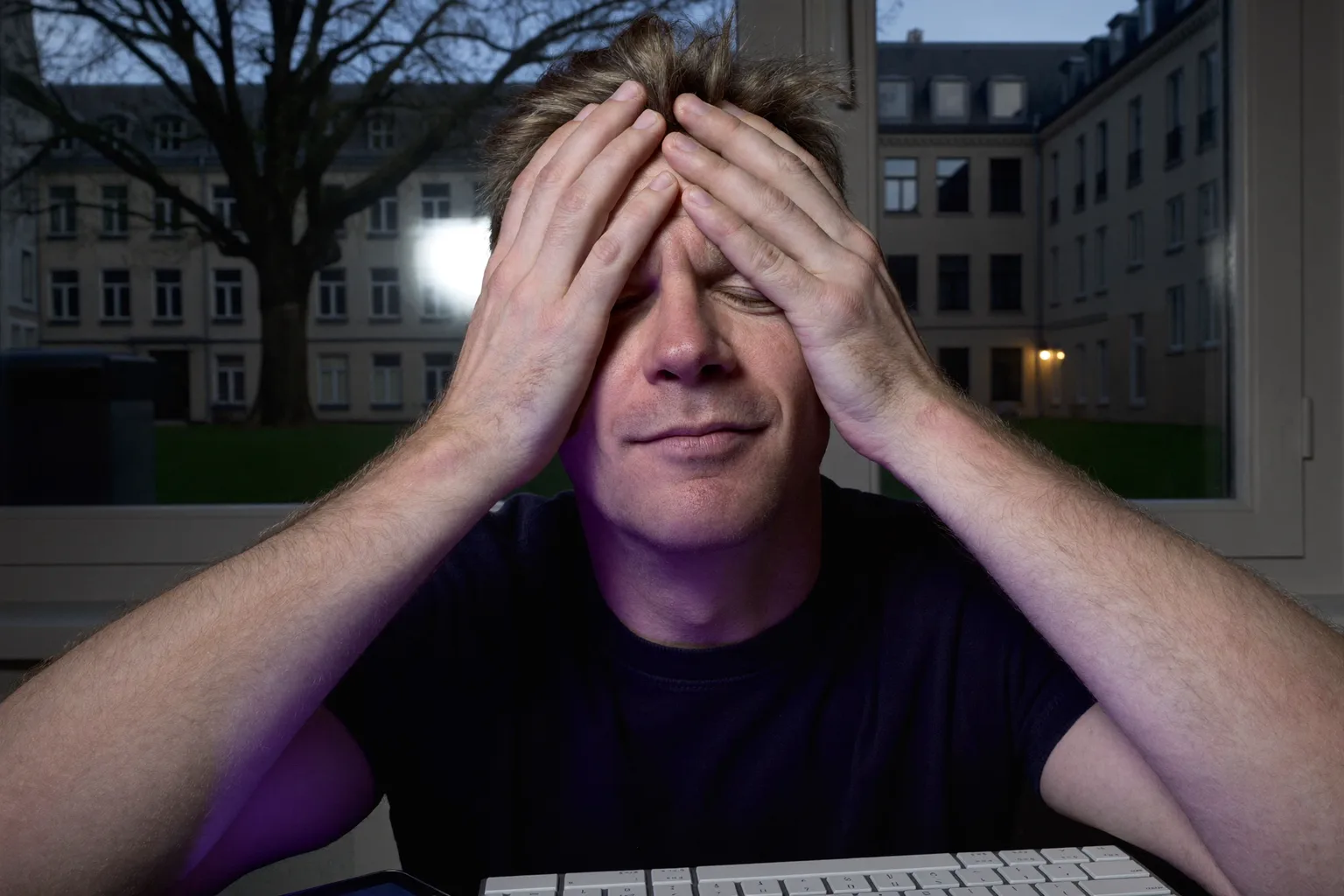 Ein Mann am Schreibtisch in einem abgedunkelten Raum presst die Handflächen auf die geschlossenen Augen, um seine müden, trockenen Augen zu entspannen, während sein Gesicht vom Laptop-Bildschirm beleuchtet wird