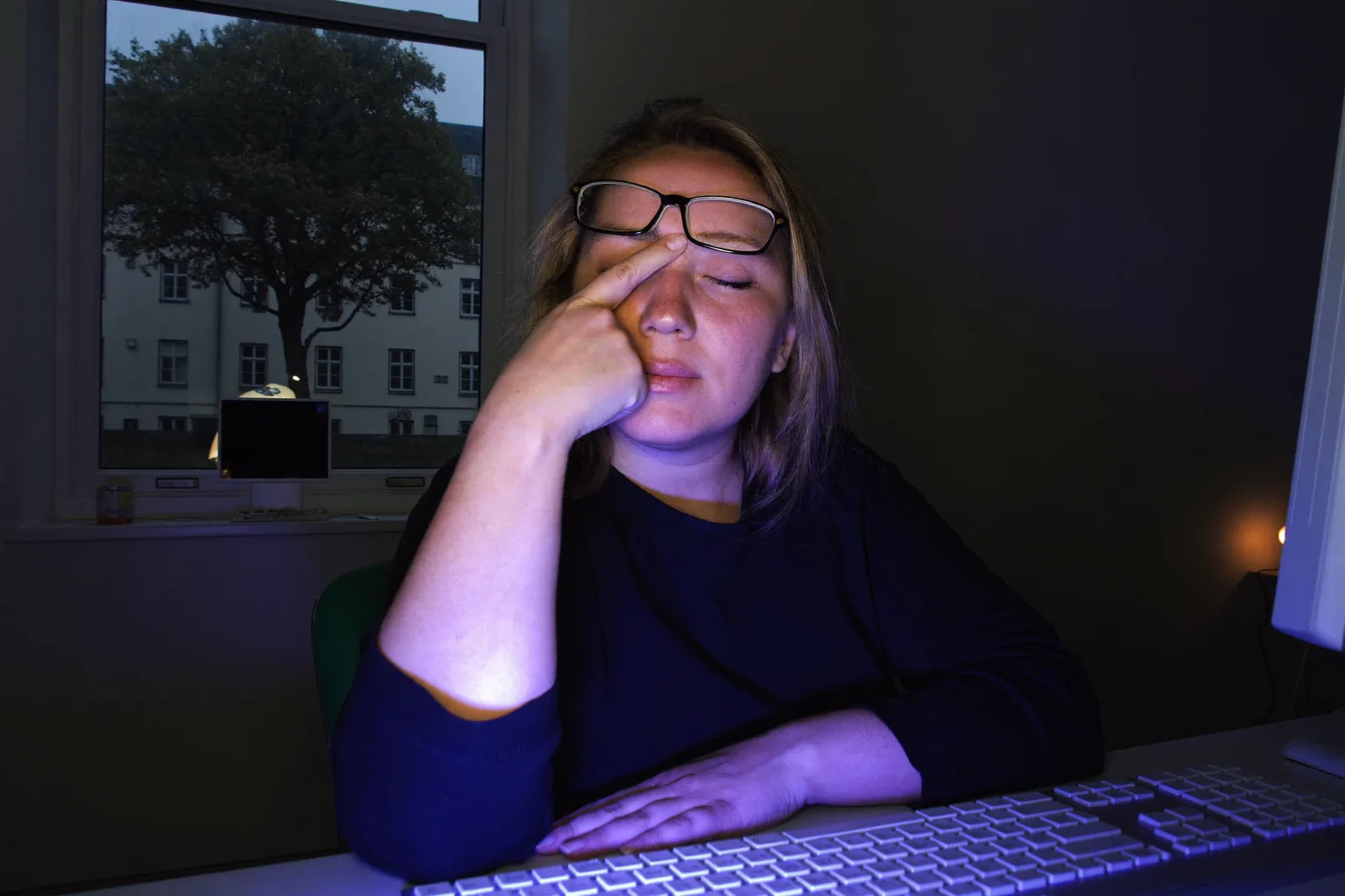 Eine Frau an einem Schreibtisch in einem dunklen Raum, die Augen geschlossen, die Brille auf die Stirn geschoben, sie drückt sich den Nasenrücken oberhalb der Rückseite ihres Monitors, das Gesicht kalt blau vom Bildschirmlicht beleuchtet