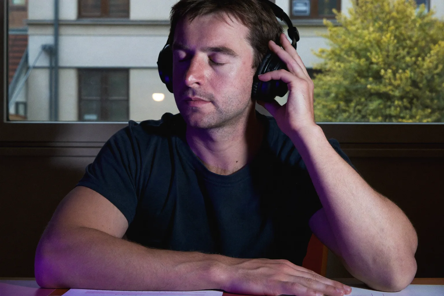Eine Person am Schreibtisch trägt Over-Ear-Kopfhörer, die Augen geschlossen, eine Hand auf der Ohrmuschel, vertieft in eine Arbeitsphase mit laufender Musik