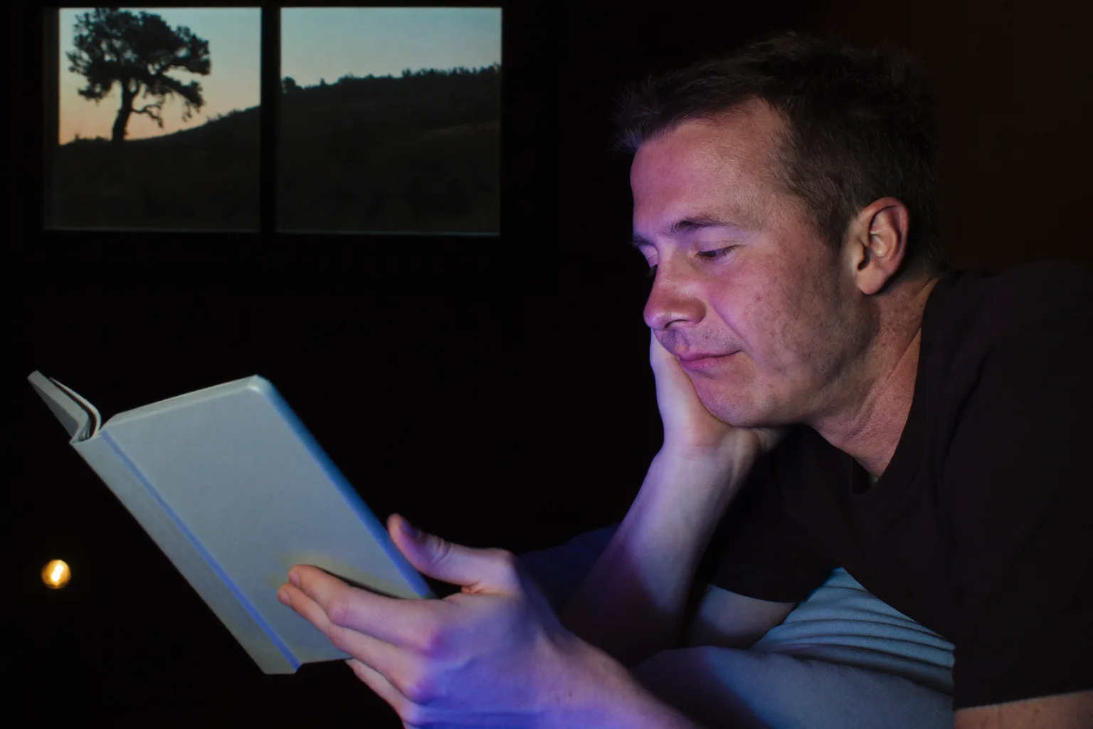 A man reading from an e-reader in bed late at night, its page a dim glow that barely reaches his face, while on the nightstand beside him a phone lying face up floods the second lampshade with hard cold blue light