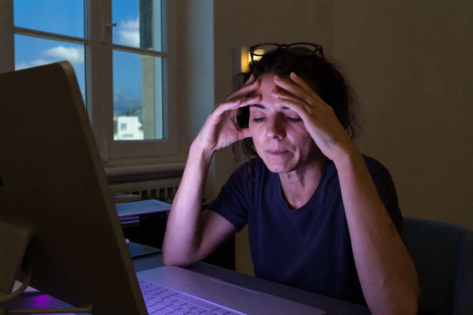 Μια γυναίκα σε ένα γραφείο μέσα σε σκοτεινό δωμάτιο, με τα μάτια κλειστά και τα γυαλιά της σηκωμένα πάνω στο μέτωπό της, πιέζει τη ρίζα της μύτης της πίσω από την οθόνη της, με το πρόσωπό της φωτισμένο σε ψυχρό μπλε από την οθόνη