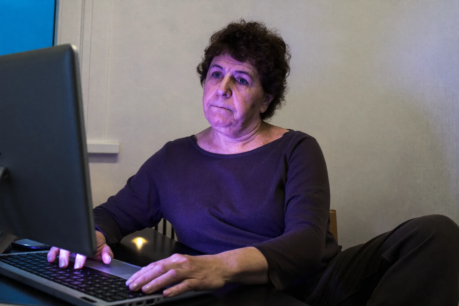 Ένα άτομο καθισμένο σε γραφείο στο σπίτι αργά το βράδυ, κοιτάζοντας πέρα από την οθόνη του laptop με τα χέρια ακόμα στο πληκτρολόγιο, δείχνοντας την επίπεδη εξάντληση της επαγγελματικής εξουθένωσης στη δουλειά