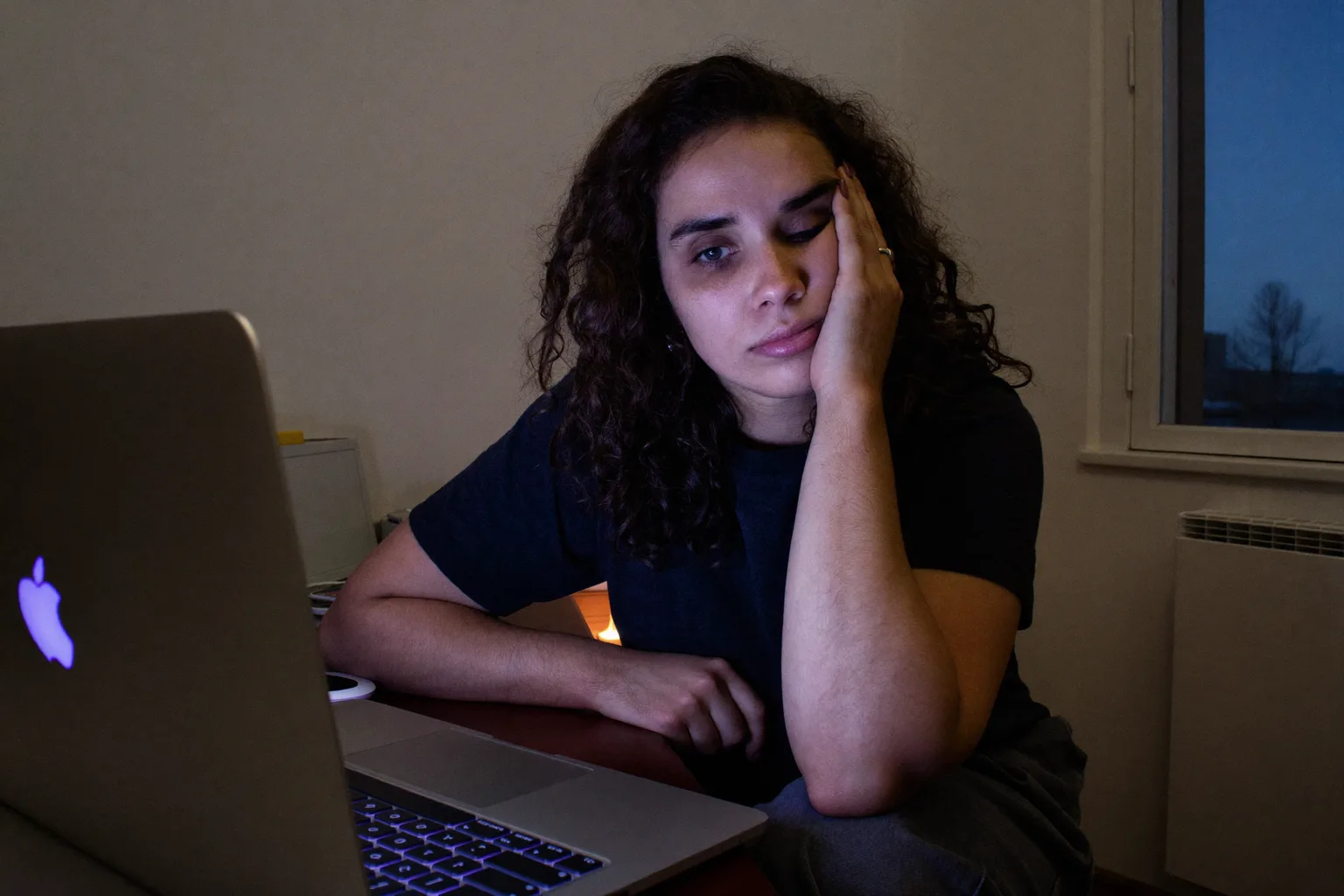 Μια κουρασμένη γυναίκα μπροστά σε λάπτοπ αργά το απόγευμα, με το κεφάλι ακουμπισμένο στο χέρι της, με βαριά, ξηρά μάτια και μαύρους κύκλους σε ένα υποφωτισμένο δωμάτιο