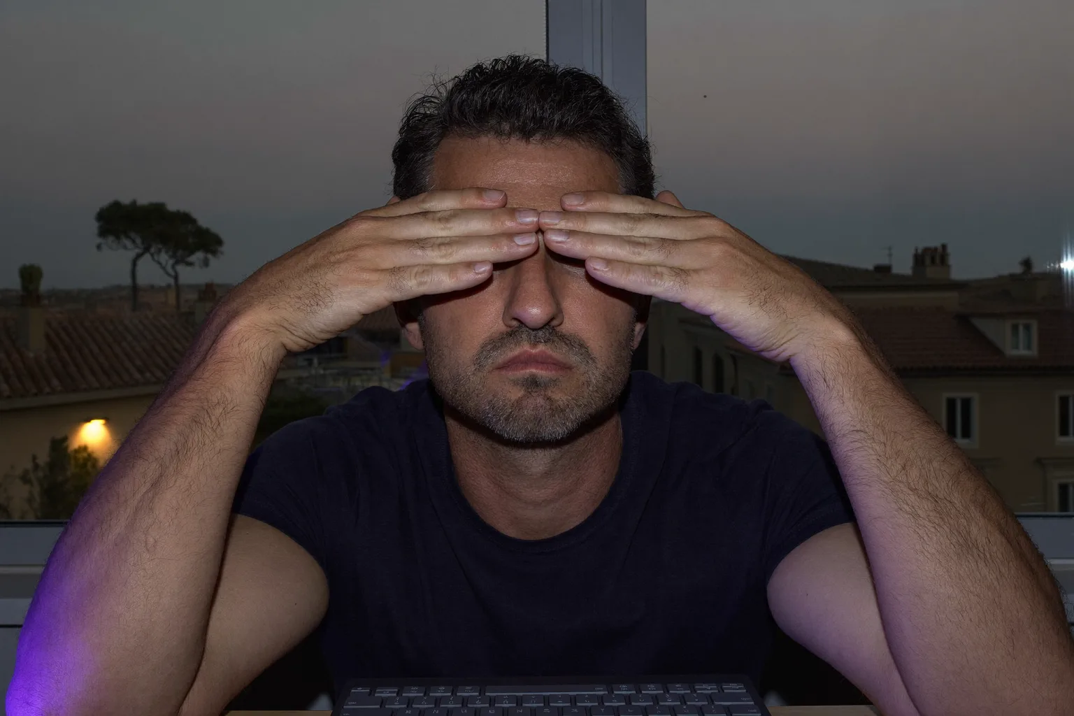 Un hombre sentado a su escritorio en una habitación en penumbra, con las palmas de las manos apoyadas sobre los ojos cerrados para aliviar la vista cansada, con el rostro iluminado por la pantalla del portátil