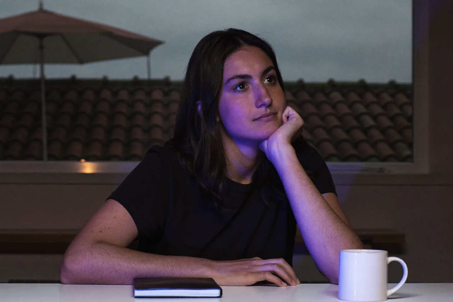 Un estudiante en un escritorio ya entrada la noche, con la barbilla apoyada en una mano abierta, el rostro iluminado de lado por el resplandor de la pantalla, la mirada perdida lejos del trabajo y fuera del encuadre