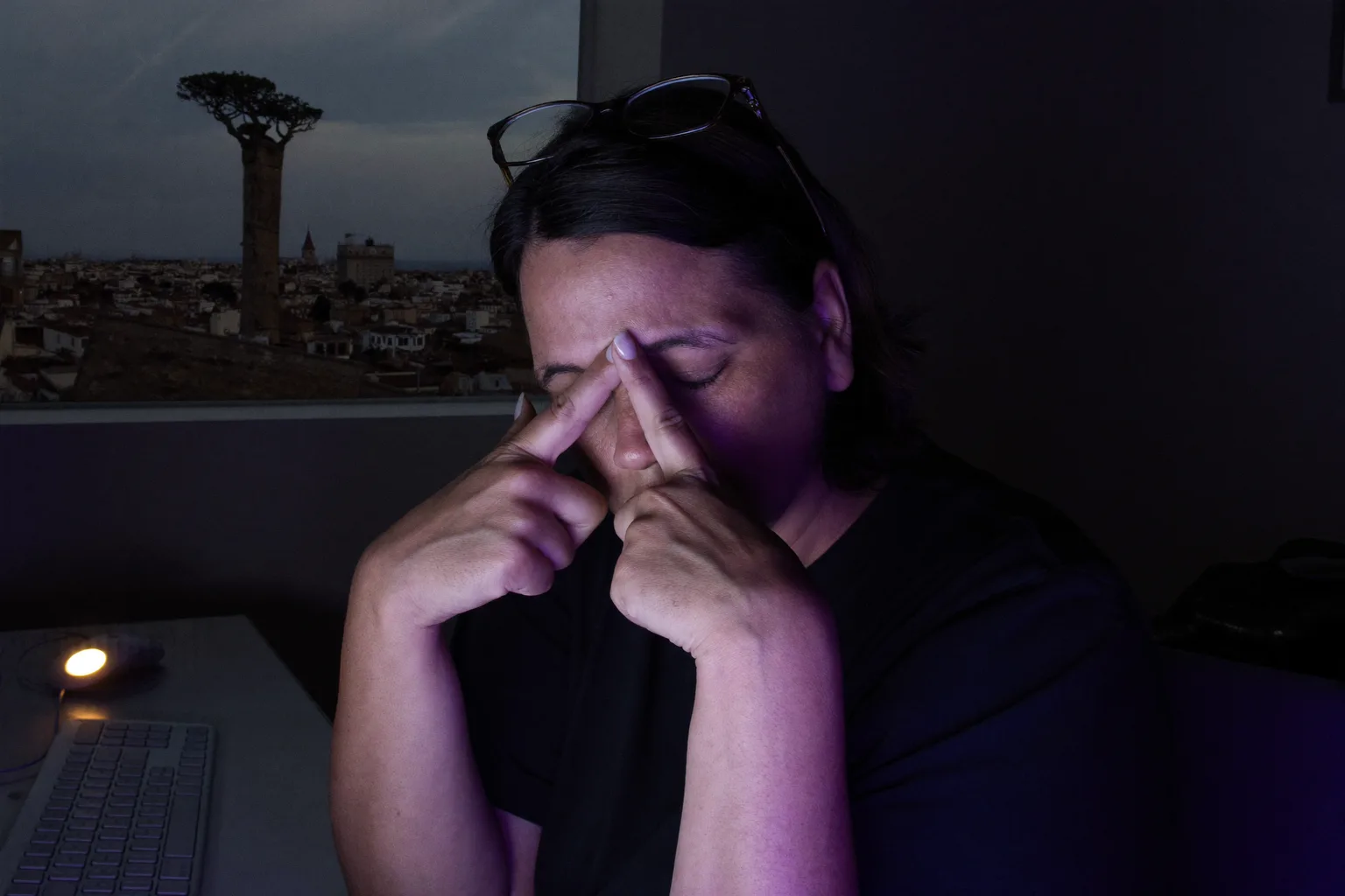 Una mujer en un escritorio, en una habitación oscura, con los ojos cerrados y las gafas subidas a la frente, apretándose el puente de la nariz por encima de su monitor, con el rostro iluminado por una luz azulada y fría de la pantalla