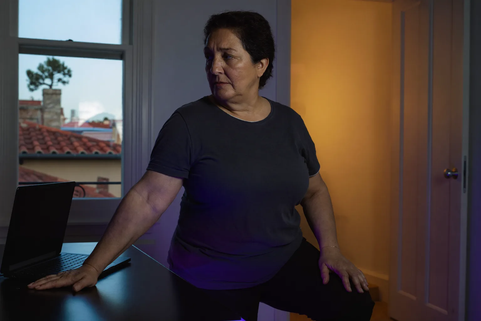 Un portátil cerrado sobre un escritorio en casa por la noche, con la luz de la habitación encendida y la persona alejándose, mostrando el final de una jornada de trabajo desde casa