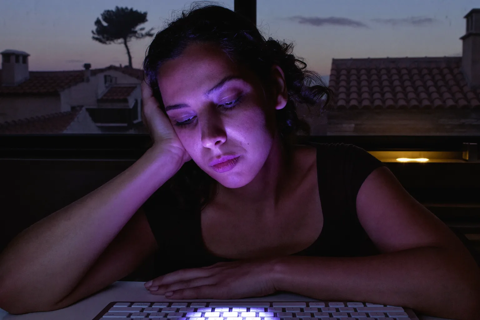 Una mujer cansada frente al portátil al final del día, con la cabeza apoyada en la mano, ojos pesados y secos y ojeras, en una habitación con poca luz