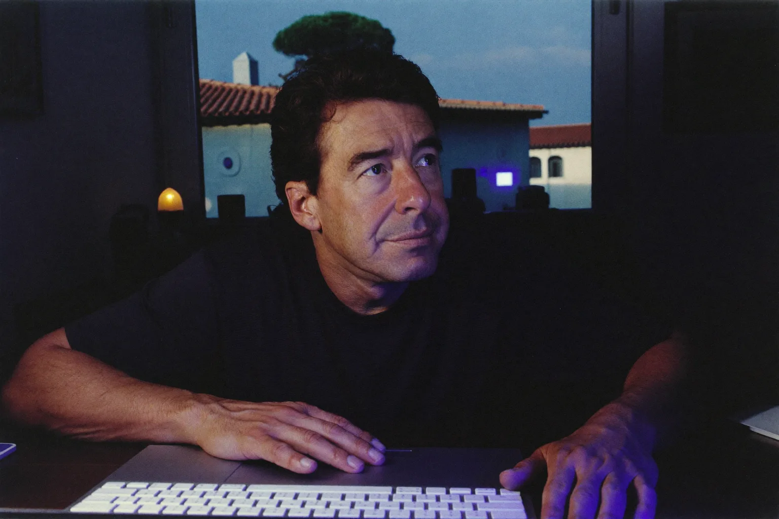 Una persona sentada en un escritorio en casa, tarde por la noche, con la mirada perdida más allá de la pantalla del portátil y las manos aún sobre el teclado, mostrando el agotamiento plano del burnout laboral
