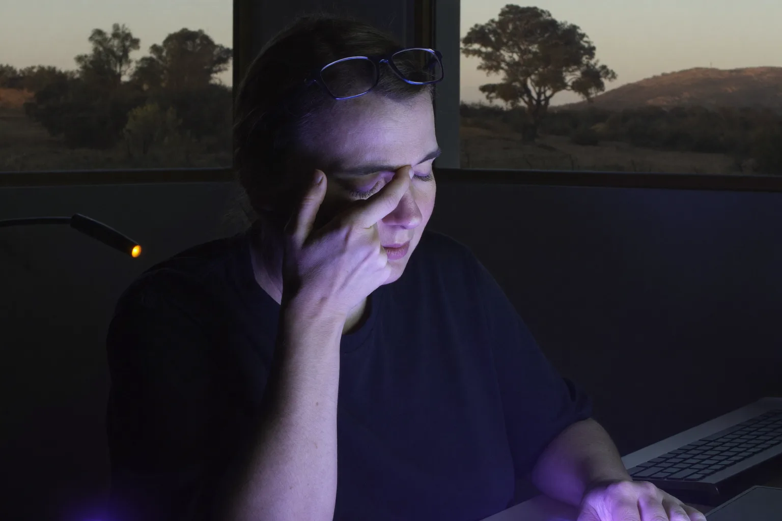 A woman at a desk in a dark room, eyes closed and her glasses pushed up on her forehead, pinching the bridge of her nose above the back of her monitor, her face lit cold blue by the screen