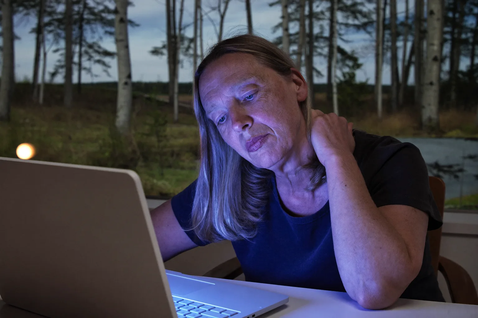 Työpisteellä istuva henkilö kurottaa kättään taakse hieromaan niskansa tyveä kannettavan tietokoneen edessä, mikä kuvastaa pitkän tietokonepäivän jäykkää ja särkevää niskaa