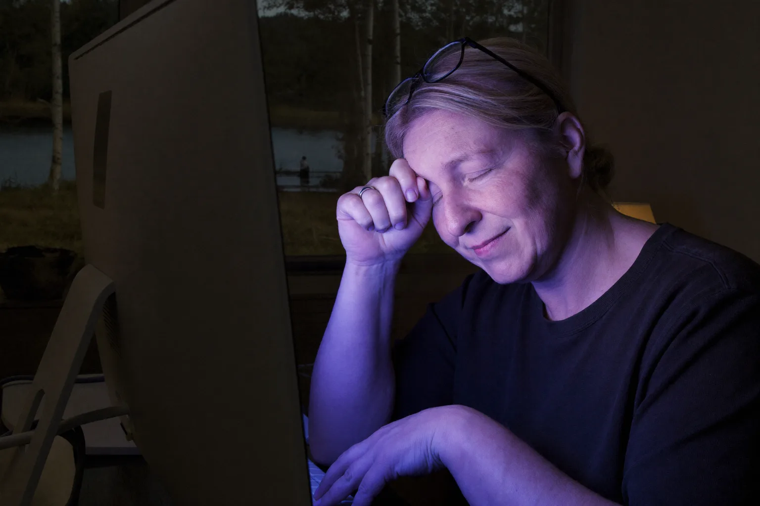 Nainen työpöydän ääressä pimeässä huoneessa, silmät kiinni ja silmälasit työnnettynä otsalle, puristaen nenänvarttaan näytön takaa, kasvot valaistuina näytön kylmän sinisellä valolla