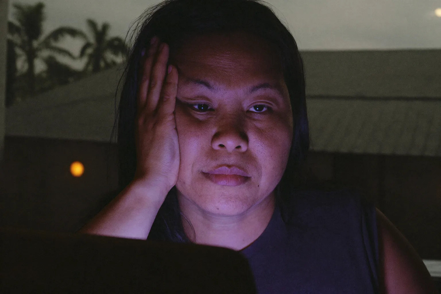Isang pagod na babae sa harap ng laptop sa gabi, ang ulo niya ay nakasandal sa kamay, may mabigat at tuyong mata at maitim na paligid ng mata sa isang madilim na kwarto