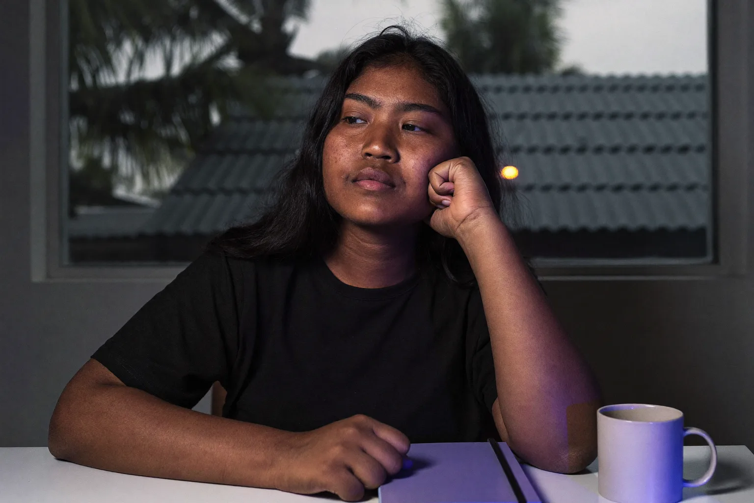 Isang estudyanteng nasa desk nang gabing-gabi na, may baba na nakasandal sa isang bukas na palad, mukhang naiilawan sa gilid ng liwanag ng screen, tingin na naligaw palayo sa ginagawa at palabas ng frame