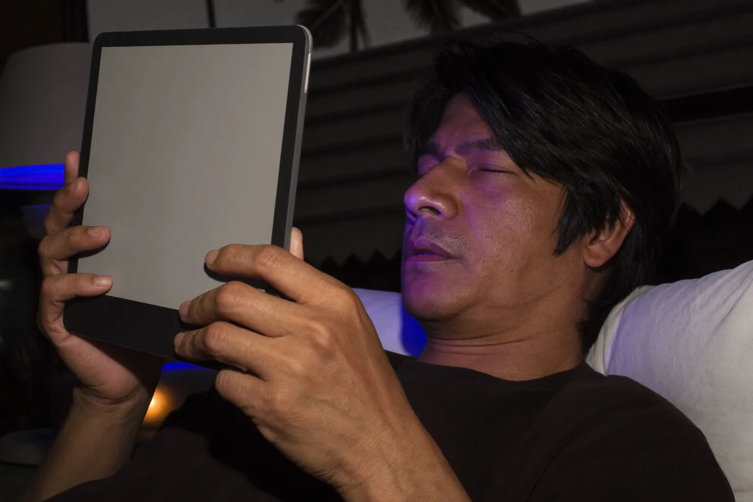 Isang lalaking nagbabasa gamit ang e-reader habang nasa kama, gabing-gabi na, ang pahina ay may mahinang liwanag na halos hindi na umaabot sa mukha niya, habang sa nightstand sa tabi niya, ang phone na nakatihaya ay pumapaligid sa pangalawang lampshade ng matigas at malamig na blue light