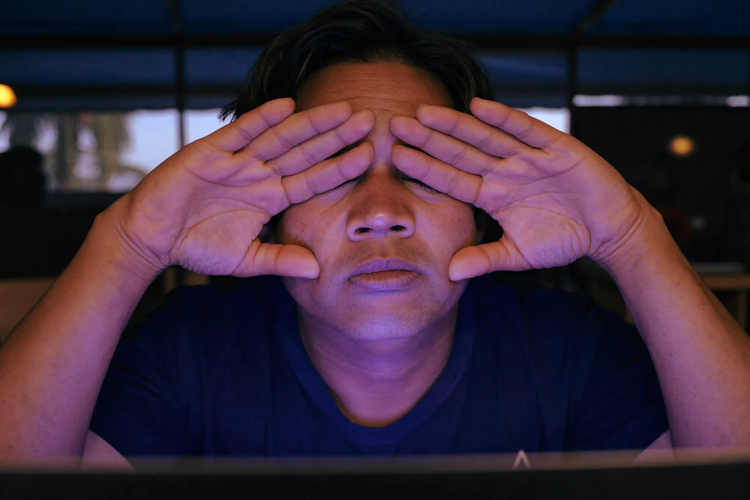 Isang lalaking nakaupo sa desk sa madilim na kwarto, itinatakip ang kanyang mga palad sa nakapikit na mata para mapawi ang pagod ng mata, ang mukha niya ay naiilawan ng liwanag mula sa laptop screen