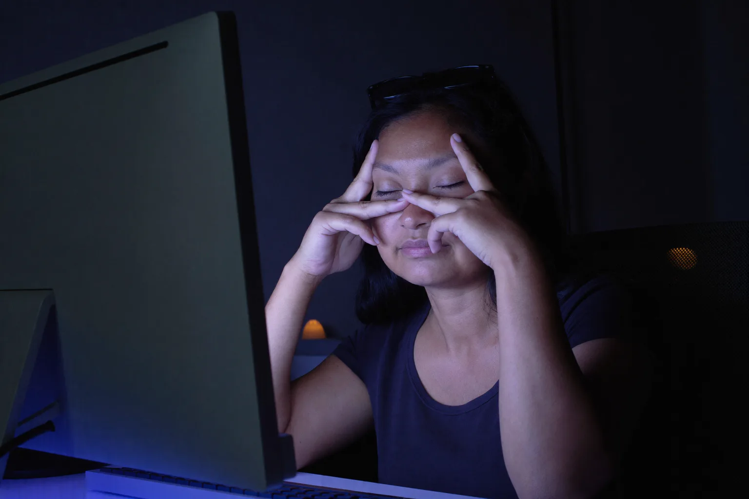 Isang babae sa harap ng desk sa madilim na kwarto, nakapikit ang mga mata at nakasampay ang salamin sa noo, pinipisil ang gitna ng ilong sa itaas ng likod ng kanyang monitor, ang mukha niya ay may malamig na asul na liwanag mula sa screen