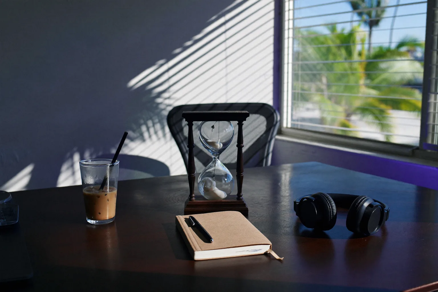 A bright Florida home office abandoned for a break: closed laptop, iced coffee sweating on the desk, palm shadows on the wall