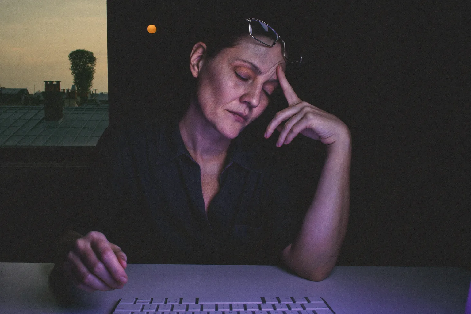 Une femme à son bureau dans une pièce sombre, les yeux fermés et les lunettes remontées sur le front, se pinçant l’arête du nez derrière son écran, le visage éclairé d’une lumière bleue froide