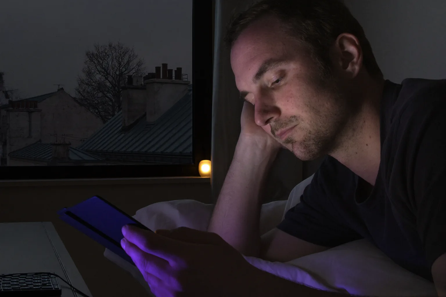 Un homme qui lit sur une liseuse au lit tard le soir, la page diffusant une lueur si faible qu’elle atteint à peine son visage, tandis que sur la table de nuit à côté de lui, un téléphone posé écran vers le haut inonde le second abat-jour d’une lumière bleue dure et froide
