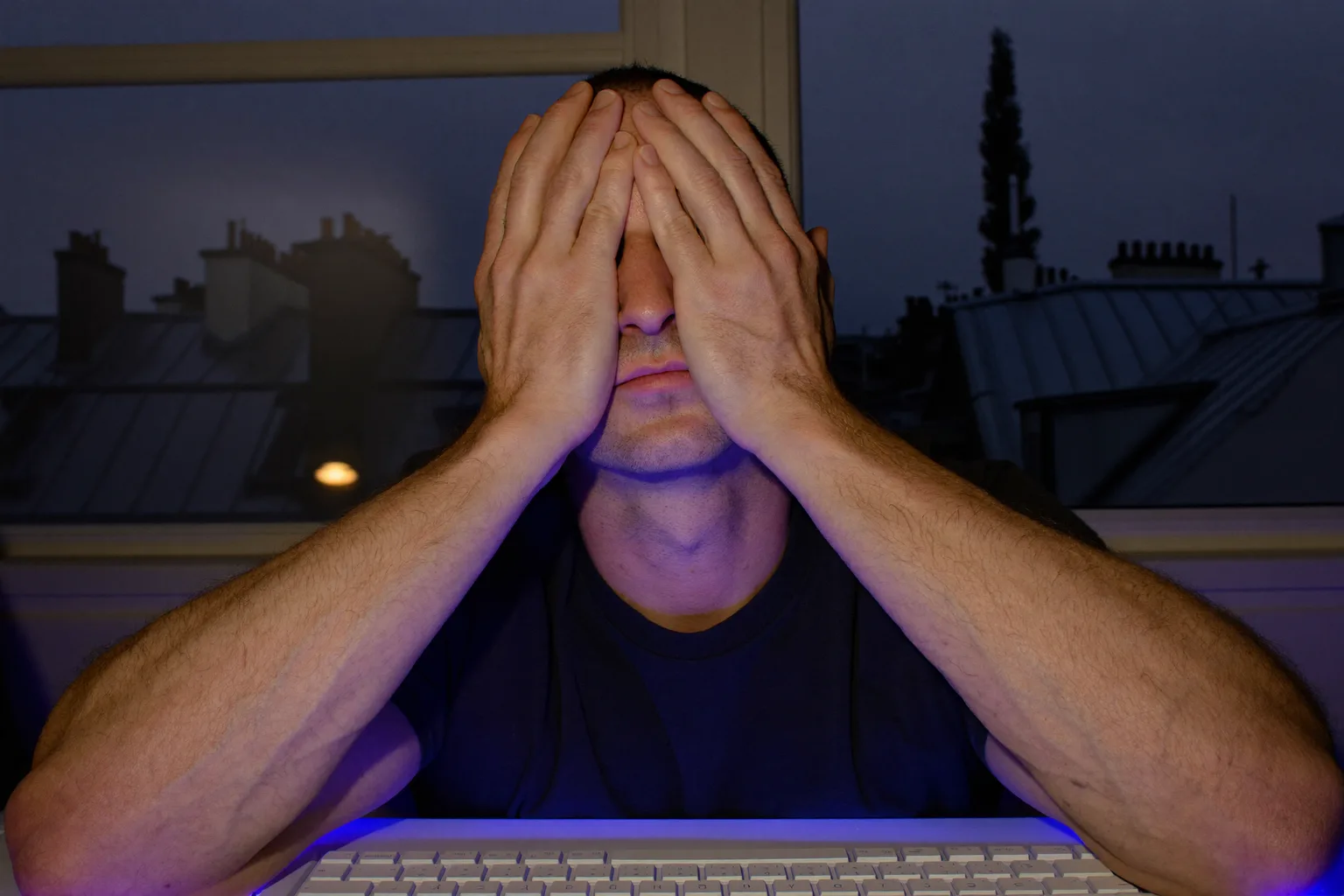 Un homme à son bureau dans une pièce sombre, les paumes posées sur ses yeux fermés pour soulager sa fatigue oculaire, le visage éclairé par l’écran de son ordinateur portable
