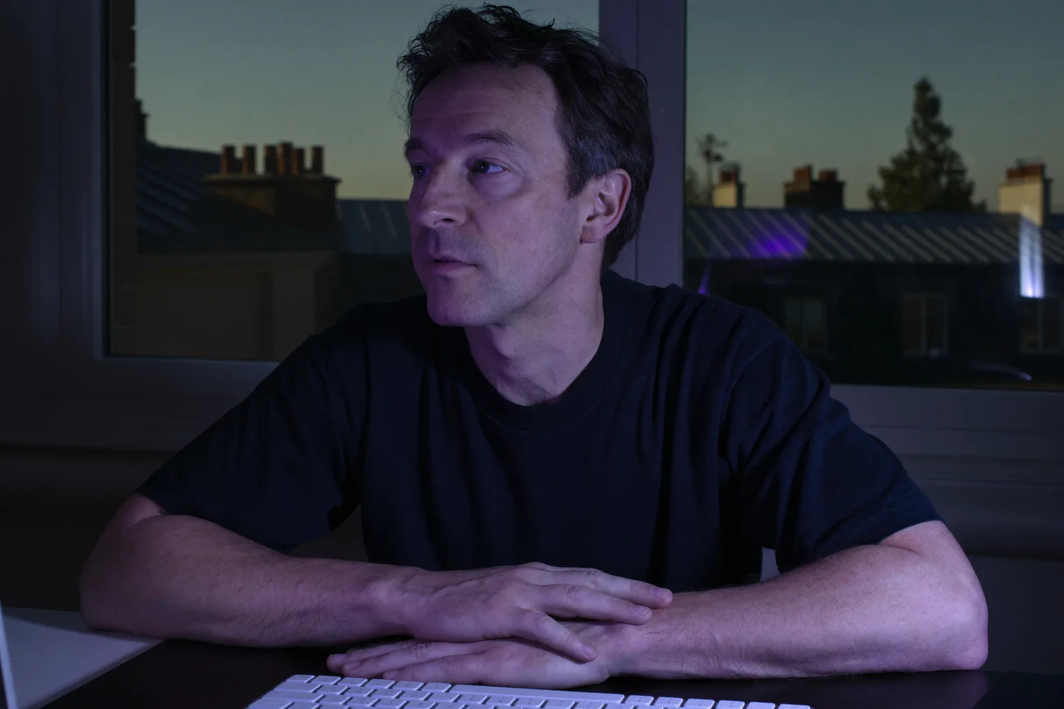 Une personne assise à son bureau à la maison tard le soir, le regard perdu au-delà de l’écran de l’ordinateur portable, les mains encore sur le clavier, l’épuisement plat du burn out au travail