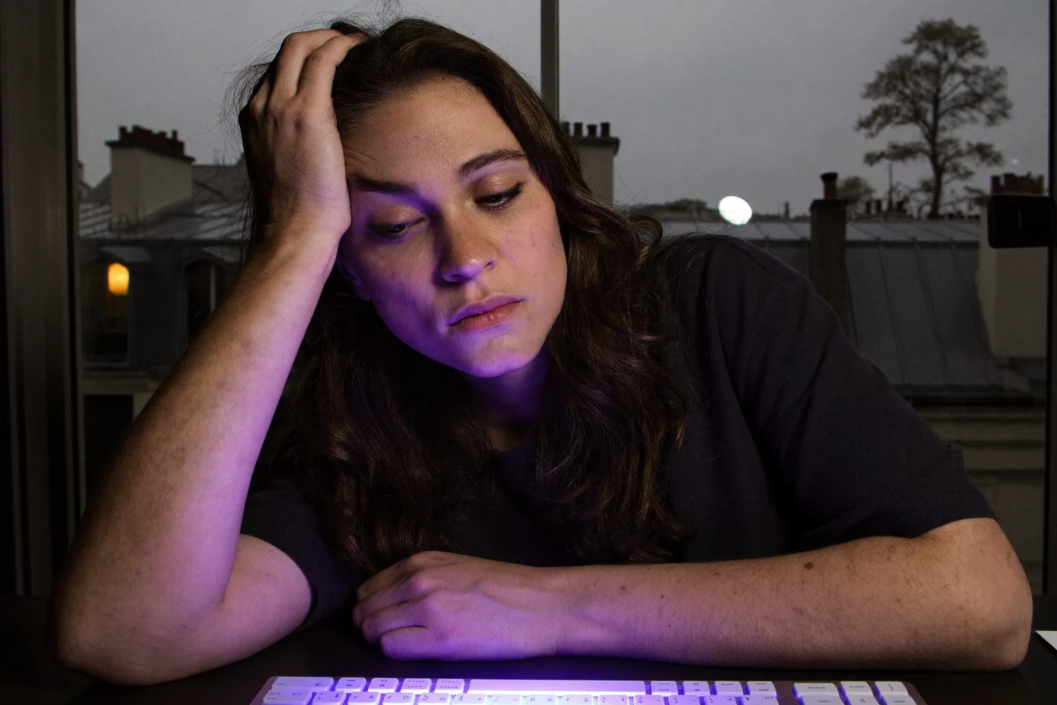 Une femme fatiguée devant un ordinateur portable en fin de journée, la tête posée sur la main, les yeux lourds et secs et des cernes, dans une pièce faiblement éclairée