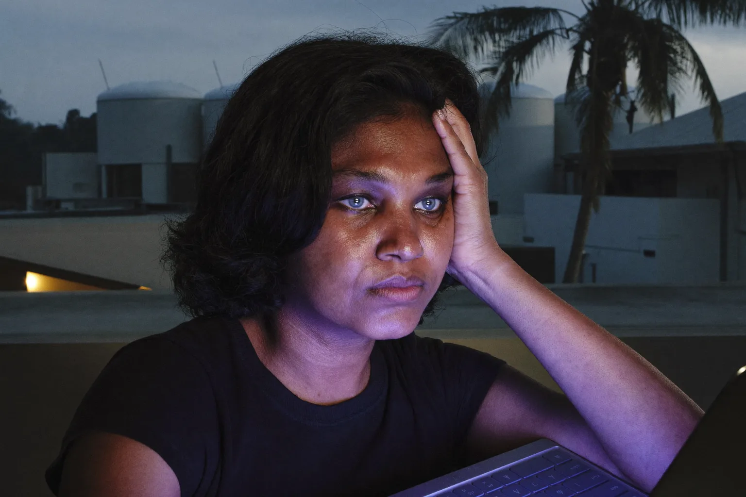A tired woman at a laptop late in the day, resting her head on her hand, with heavy, dry eyes and dark circles in a dimly lit room