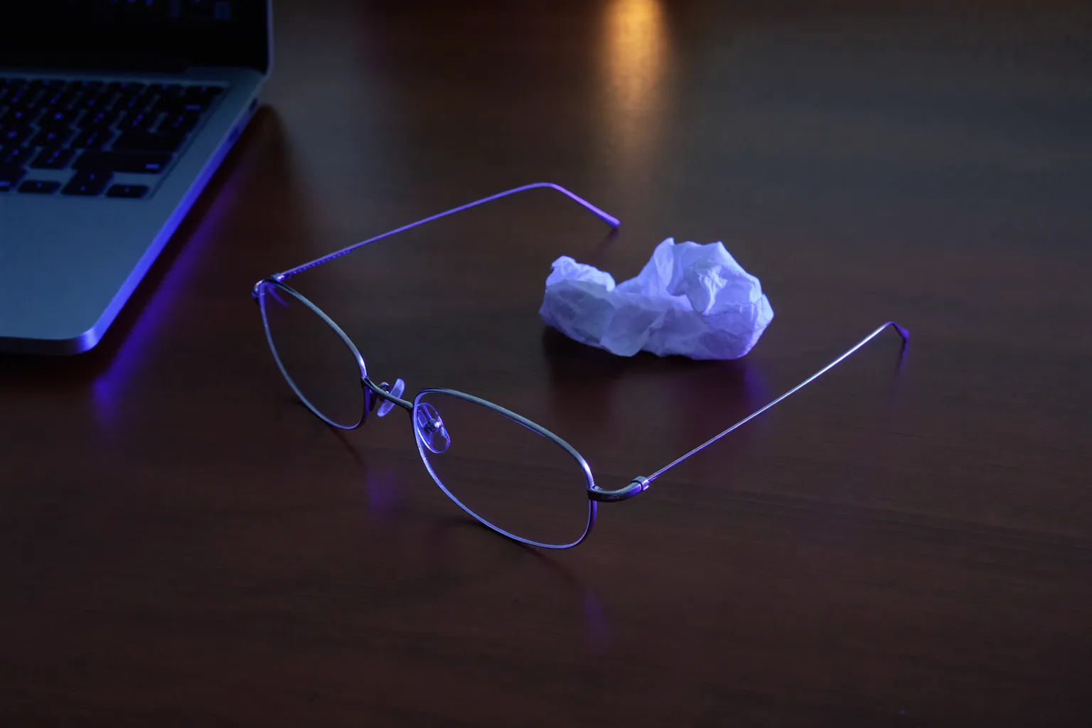 A pair of reading glasses folded on a dark desk at dusk beside a crumpled tissue, next to a laptop whose screen is out of frame