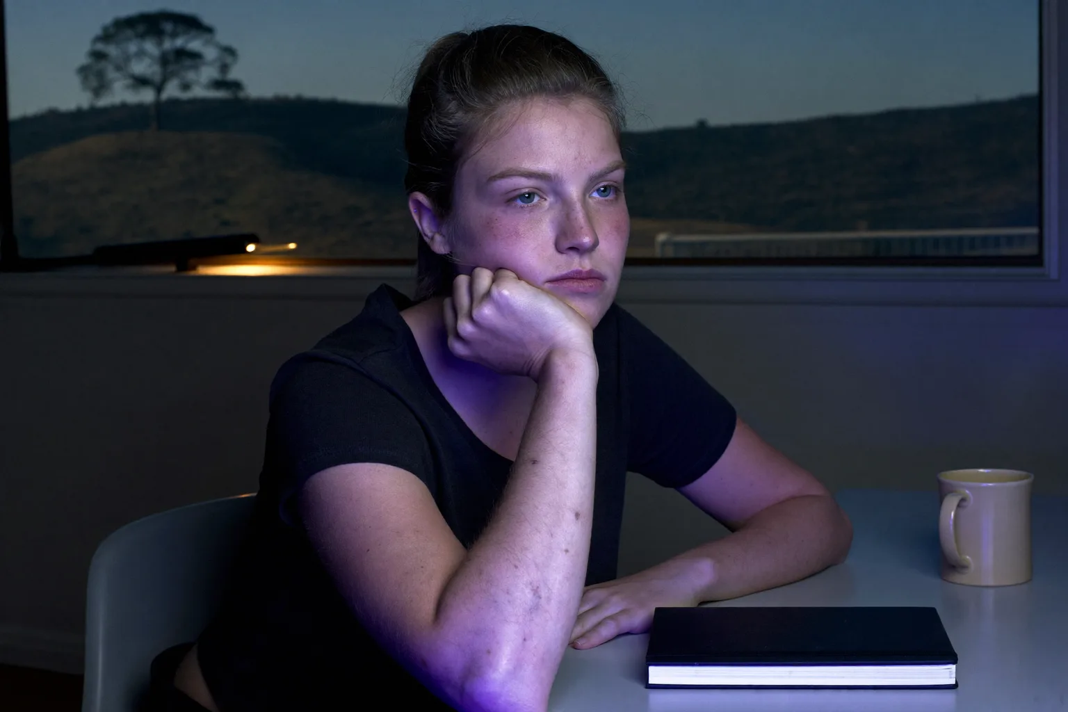 A student at a desk late in the evening, chin resting in one open palm, face lit from the side by screen glow, gaze drifted away from the work and off the frame