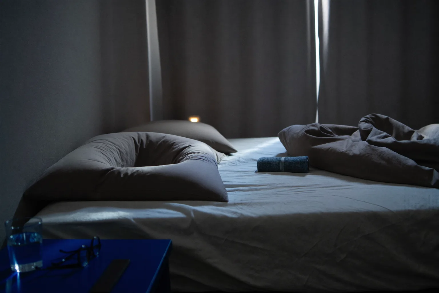 An empty unmade bed at first light, one pillow still holding the deep hollow left by a head, a rolled towel on the sheet, and a glass of water beside a folded pair of glasses on the bedside table