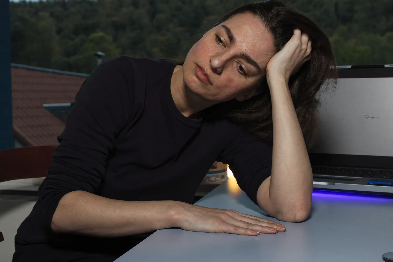 Osoba za kućnim radnim stolom kasno navečer, glave naslonjene na jednu ruku s prstima u kosi, oborenog i nefokusiranog pogleda, dok druga podlaktica mirno leži na praznom stolu