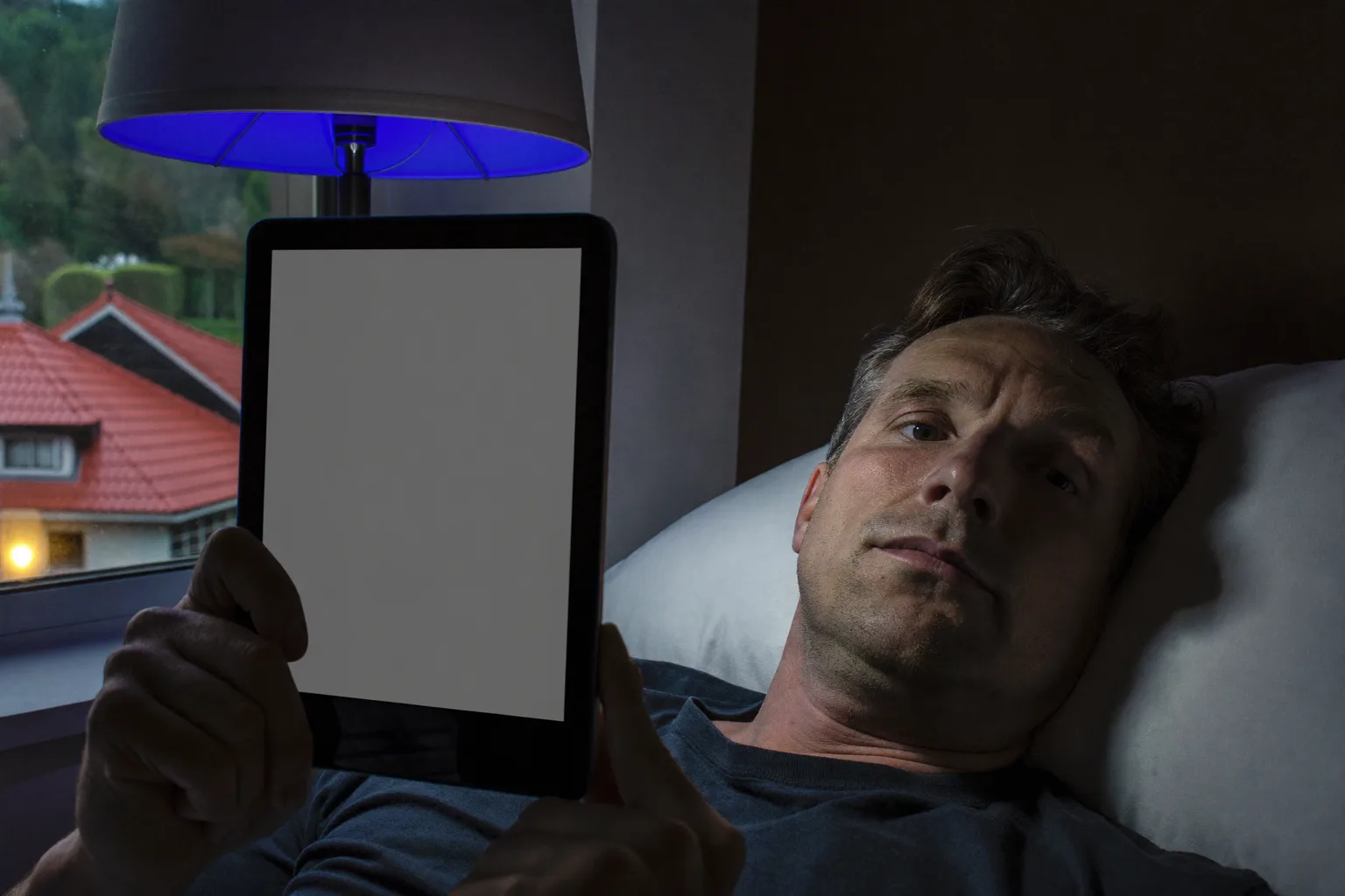 Muškarac čita s e-čitača u krevetu kasno navečer, a njegova stranica daje slab sjaj koji jedva doseže njegovo lice, dok telefon okrenut ekranom prema gore na noćnom ormariću pored njega oblijeva susjedno sjenilo lampe oštrim, hladnim plavim svjetlom