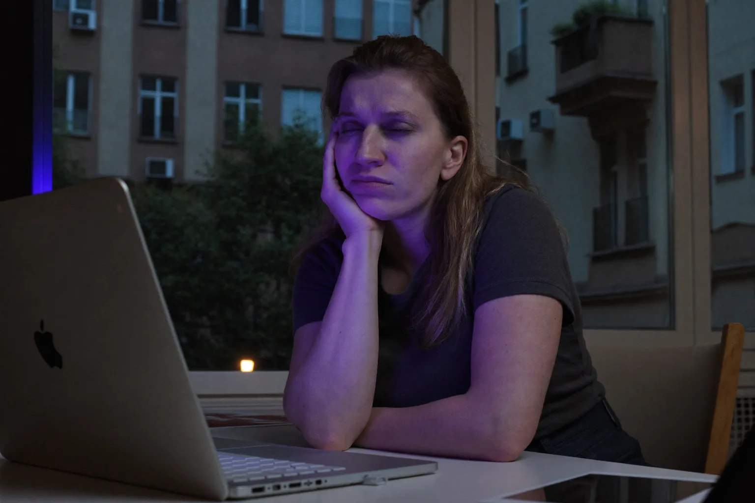 Egy fáradt nő a laptopnál a nap végén, kezére támasztva a fejét, nehéz, száraz szemekkel és sötét karikákkal, egy halványan megvilágított szobában