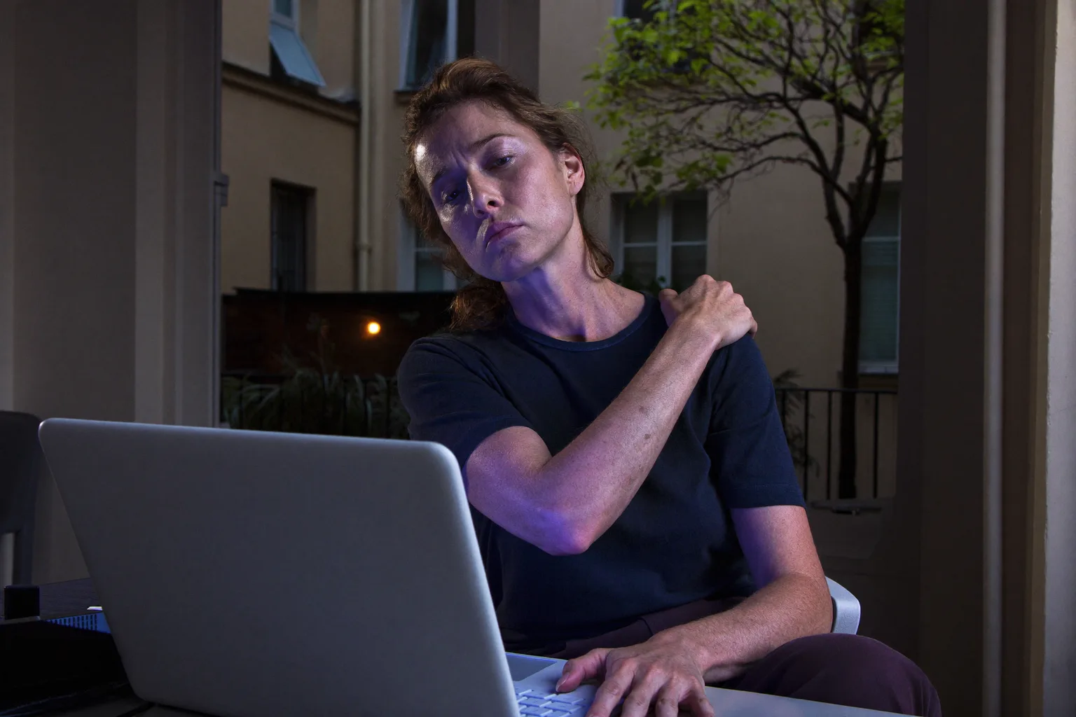 Egy ember az íróasztalánál hátranyúl, hogy megdörzsölje a nyaka alját egy laptop előtt, ami egy hosszú számítógépes nap merev, fájó nyakát mutatja